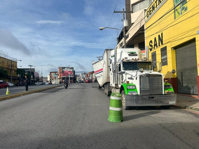 First image displays a street scene in an urban area with multiple large white semi-trucks parked along the roadside near yellow buildings marked with SAN and GENTA signs, traffic cones in green and white, a motorcycle, cars, and pedestrians under a partly cloudy sky with streetlights and billboards visible. Second image shows a yellow overpass bridge with a large overturned or damaged truck trailer wedged against it, another truck nearby, and construction barriers with yellow and black stripes on a curved road section. Third image depicts a white truck trailer labeled DH with number 02, severely damaged and tilted inside a dimly lit underpass area, cargo spilled out, wheels askew on wet pavement.