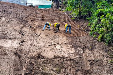 Three images show construction workers in blue jeans and yellow safety vests bending over to install wire mesh on a steep earthen slope reinforced with metal rods, surrounded by green vegetation and temporary structures like metal sheds and green netting in a roadside setting in Guatemala.