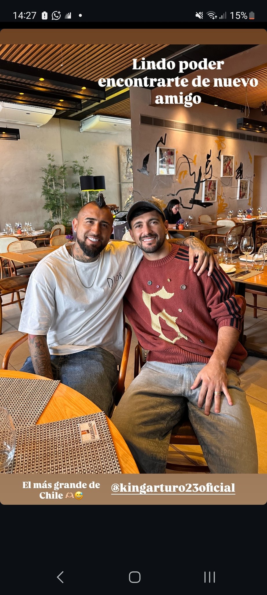 Two men with beards sit closely at a wooden restaurant table, one wearing a white t-shirt and the other a red Adidas hoodie, both smiling at the camera with arms around each other, surrounded by wooden chairs, plants, and wall art in a modern indoor dining space.
