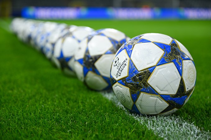 A row of white UEFA Champions League soccer balls with blue and gold designs aligned on green grass in a stadium. The balls display the UEFA Champions League logo prominently. Stadium seating and blue banners appear in the background. One ball is positioned in the foreground for emphasis.