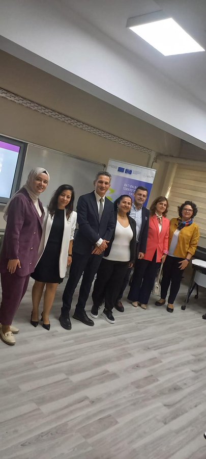 First image shows a large group of about 15-20 adults including men in suits and women in professional attire and headscarves standing in front of a yellow school building with a blue sign reading ORTAOKULU. Second image depicts six adults in business clothes including headscarves posing inside a classroom with a projector screen displaying MEB content and tables. Third image features four adults in formal wear standing on steps in front of a pink and yellow school building with a sign reading ÇANAKKALE MERKEZİ CEVAT PAŞA ORTAOKULU. Fourth image shows a classroom with around 20 students in uniforms seated at desks using laptops while a male teacher in suit stands presenting near a computer screen.