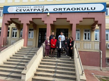 First image shows a large group of about 15-20 adults including men in suits and women in professional attire and headscarves standing in front of a yellow school building with a blue sign reading ORTAOKULU. Second image depicts six adults in business clothes including headscarves posing inside a classroom with a projector screen displaying MEB content and tables. Third image features four adults in formal wear standing on steps in front of a pink and yellow school building with a sign reading ÇANAKKALE MERKEZİ CEVAT PAŞA ORTAOKULU. Fourth image shows a classroom with around 20 students in uniforms seated at desks using laptops while a male teacher in suit stands presenting near a computer screen.