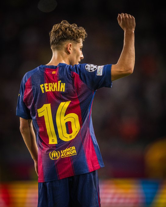Player Fermin Lopez stands on a soccer field at night, viewed from behind, wearing a navy blue and garnet red FC Barcelona jersey with number 16 on the back, UEFA Champions League sleeve patch, and sponsor logos including UNICEFs childrens fund emblem, raising his right fist in a celebratory gesture, with stadium lights and blurred crowd in the background.