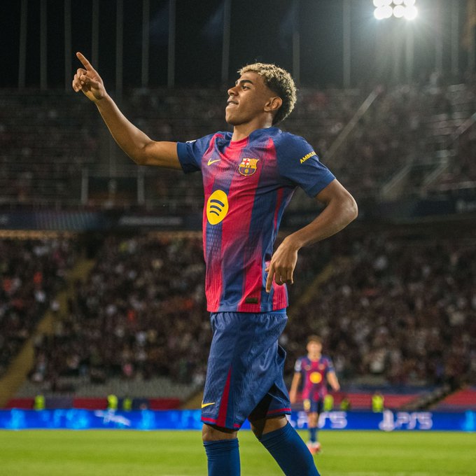 Lamine Yamal in FC Barcelona jersey points one finger skyward with a grinning expression on a vibrant green soccer field under stadium floodlights at night. He wears blue shorts and the teams blaugrana kit with Spotify sponsor patch visible. Blurred stadium seating and another player in similar uniform appear in the background with PlayStation advertising boards along the pitch edge.