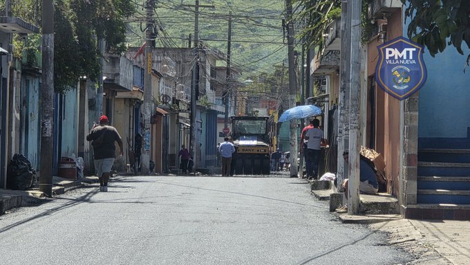 A narrow urban street lined with colorful buildings and power lines under a clear sky. Workers in casual clothing operate a yellow bulldozer-like vehicle in the center. Pedestrians including a man with an umbrella walk along the sidewalks. A blue shield with PMT_VILLANUEVA logo is visible on a wall. Vegetation and parked items like bags are present on the sides.