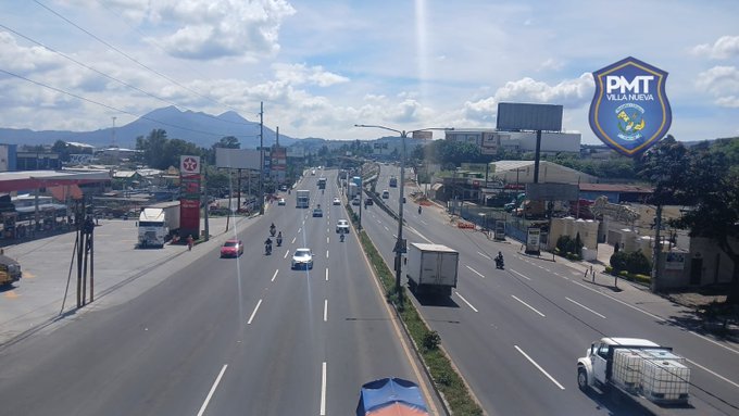 Aerial perspective of a multi-lane highway at km 15 on CA-9 showing northbound and southbound traffic with white cars, red vehicles, trucks, motorcycles, and a water tanker under clear skies with scattered clouds and green mountains in the background, billboards and roadside structures visible, PMT shield logo overlaid in the corner.