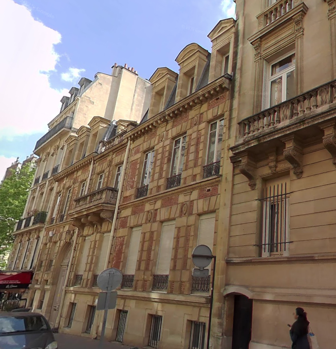 A row of historic beige stone buildings with ornate facades, arched windows, balconies, and mansard roofs under a partly cloudy blue sky. The structures feature decorative cornices, shutters, and wrought-iron details typical of 19th-century Parisian architecture. A narrow street runs in front with a small car parked nearby and a person in casual clothing walking on the sidewalk. Trees and a street sign are visible on the left side.
