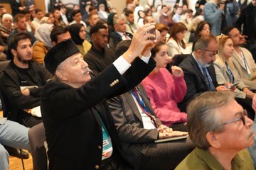 First image shows a conference room with a large screen displaying Turkish landmarks like a mosque and bridge, a speaker in a suit at a podium addressing the audience, wooden paneling, chairs, and attendees in formal attire seated and standing. Second image features a poster for Turkoloji Sempozyumu on Turkish teaching in Balkans with photos of three male speakers in suits at podiums, named Mustafa Uzbas, Cem Zorlu, Ali Gunes. Third image depicts a diverse audience at the event including men and women in traditional and modern clothing, one elderly man in a black cap taking a selfie with a phone. Fourth image shows a panel of seated men and women in business attire and headscarves around a table with water bottles, in a modern conference setting with cameras.