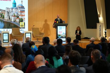 First image shows a conference room with a large screen displaying Turkish landmarks like a mosque and bridge, a speaker in a suit at a podium addressing the audience, wooden paneling, chairs, and attendees in formal attire seated and standing. Second image features a poster for Turkoloji Sempozyumu on Turkish teaching in Balkans with photos of three male speakers in suits at podiums, named Mustafa Uzbas, Cem Zorlu, Ali Gunes. Third image depicts a diverse audience at the event including men and women in traditional and modern clothing, one elderly man in a black cap taking a selfie with a phone. Fourth image shows a panel of seated men and women in business attire and headscarves around a table with water bottles, in a modern conference setting with cameras.