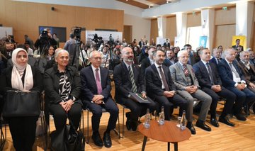 First image shows a conference room with a large screen displaying Turkish landmarks like a mosque and bridge, a speaker in a suit at a podium addressing the audience, wooden paneling, chairs, and attendees in formal attire seated and standing. Second image features a poster for Turkoloji Sempozyumu on Turkish teaching in Balkans with photos of three male speakers in suits at podiums, named Mustafa Uzbas, Cem Zorlu, Ali Gunes. Third image depicts a diverse audience at the event including men and women in traditional and modern clothing, one elderly man in a black cap taking a selfie with a phone. Fourth image shows a panel of seated men and women in business attire and headscarves around a table with water bottles, in a modern conference setting with cameras.