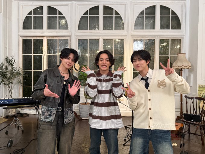 Three young men stand in a bright indoor studio with large arched windows and plants. The left man wears a dark jacket over a black shirt with hands raised. The center man in a striped shirt also raises hands excitedly. The right man in a light cardigan and tie makes a peace sign. A lamp and keyboard are visible nearby.