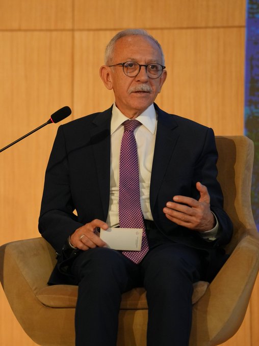 First image shows an elderly man with glasses and mustache wearing a dark suit white shirt and patterned red tie sitting in a beige armchair gesturing with his right hand while holding a white card in his left hand in front of a wooden paneled wall with a microphone stand. Second image displays a panel discussion stage with a blue banner reading 4th International Balkan Turkish Language Teaching and Turkology Symposium 2025 Panel on Migration Traces in the Balkans featuring photos of panelists Ahmet Ebilgili Prof Dr Senem Cevik Prof Leyla Sevim and Mehmet Toker seated at a table with microphones and screens in a modern auditorium. Third image captures a group of panelists including men and women in formal business attire seated on stage with a blue backdrop showing names like Ahmet Ebilgili and Leyla Sevim surrounded by audience members clapping in a conference setting.