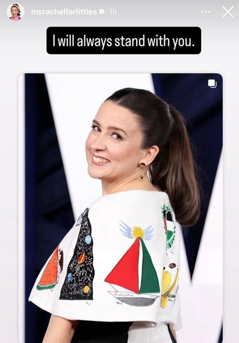 A woman with shoulder-length dark hair in a ponytail smiles while looking to the side, wearing a white cape-like top adorned with colorful childrens drawings including watermelons, stars, sailboats, birds, and flags, over a black outfit, positioned against a neutral background with subtle lighting.