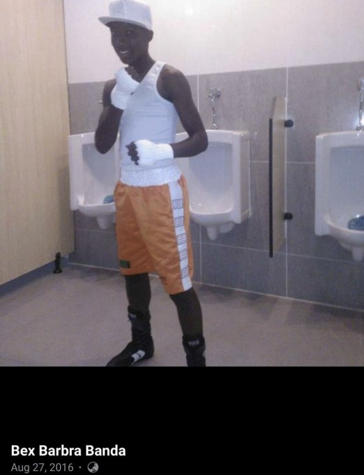 A young individual with short hair, dark skin, wearing a white tank top, orange boxing shorts with white stripes, white boxing gloves, black sneakers, and a white cap, posing confidently with fists up in a tiled bathroom featuring multiple white urinals on a gray wall background.