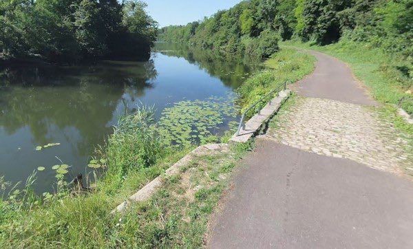 A serene riverside path along a calm body of water with lush green trees and vegetation on both sides. The water surface shows reflections and patches of green lily pads. A paved walkway runs parallel to the riverbank, with a stone or cobblestone ramp leading down to the edge. The scene appears peaceful under clear skies, capturing a natural outdoor setting near the Saône river where the body was discovered.