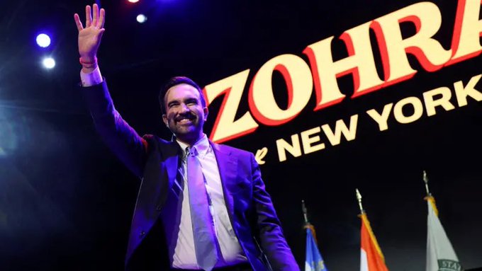 A man in a dark suit with a light blue tie stands on a stage raising his right hand in a wave while smiling, positioned in front of large illuminated text reading Zohran for New York with colorful stage lights and several flags including American, orange, and blue ones visible in the background.