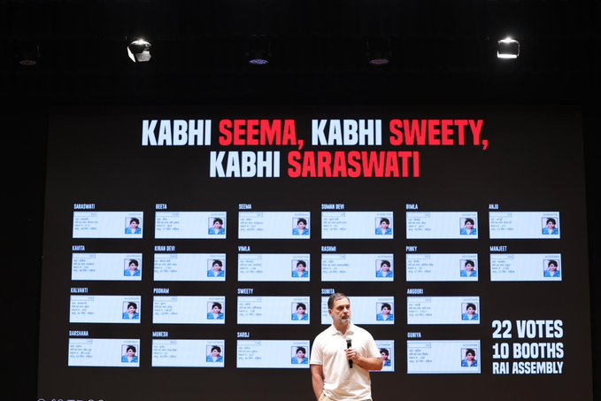 A man in a white shirt and jeans stands on a stage holding a microphone, speaking at a political rally. Behind him is a large dark screen displaying the text Kabhi Seema Kabhi Sweety Kabhi Saraswati in red and white, with multiple small photos of women arranged in a grid below it. The screen also shows 10 Booths 22 Votes Raebareli Assembly at the bottom. Stage lights illuminate the area.