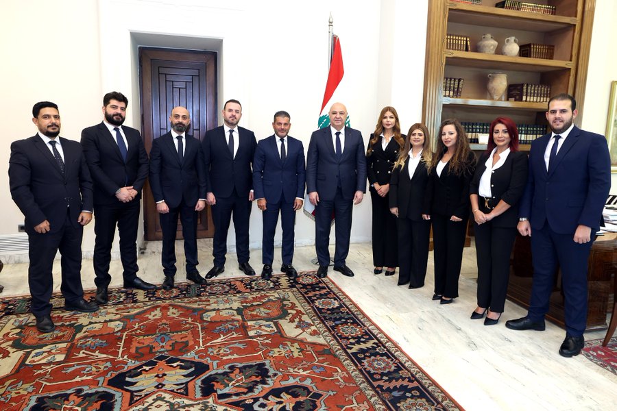 Group of ten people including men in dark suits and two women in formal attire one in gray suit and one in white blouse stand in two rows in an elegant office with wooden bookshelves vases on shelves a large wooden door behind them a Lebanese flag on a pole to the right a large red and gold patterned rug on the wooden floor and a wooden side table nearby