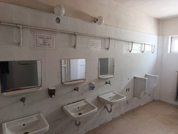 First image shows a school classroom wall with blue barred window, bulletin board displaying orange and green posters on hygiene and class schedules, clock, and two labeled waste bins one gray for general waste and one blue for recyclables on light blue tiled floor. Second image depicts a white tiled wall with a printed attendance or log sheet pinned up and a poster warning against drug use featuring illustrative graphics. Third image displays a school restroom with white tiled walls, multiple white sinks with faucets soap dispensers, mirrors, urinals, and overhead lights. Fourth image portrays an outdoor school courtyard with paved ground, basketball hoop, colorful playground slide, pine trees, Turkish flag on beige building with entrance sign, and fence.