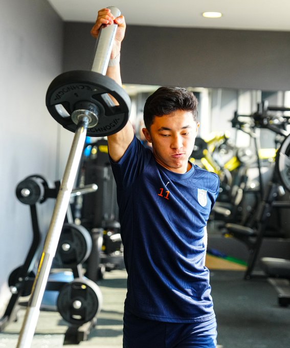 First image shows a young Asian male athlete in a navy blue team jersey numbered 11 lifting a barbell with weight plates in a modern gym surrounded by equipment and mirrors. Second image depicts a curly-haired male athlete in navy blue long-sleeve shirt performing a deadlift with a barbell and plates in the same gym setting with other players visible. Third image features a dreadlocked Black male athlete in a sleeveless navy blue jersey numbered 23 doing cable machine pulls with resistance bands attached. Fourth image captures a bald male athlete in navy blue long-sleeve top leading a group exercise on a step platform with teammates participating in a fitness routine amid gym apparatus like exercise balls and weights.
