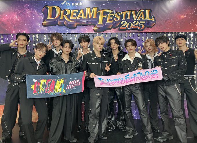 Group of 11 young men in black leather jackets and pants posing energetically in front of a starry backdrop with Dream Festival 2025 banner and TV Asahi logo. They hold colorful banners reading Dreams Festival and Dream Festival. The members have styled hair and accessories, standing on a stage-like area with event decorations.