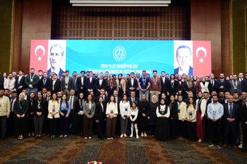 Large conference hall with blue and red banners displaying Turkish flags and text for 2024 YLSY Bursiyerleri Vizyon ve Farkındalık Eğitimi Programı featuring large portraits of Yusuf Tekin at podium with audience seated below stage with microphones and decorative elements second image shows elegant hall with chandeliers wooden panels Turkish flags and suited men including speaker at white podium with flower arrangements third image depicts auditorium with green walls large screen showing event logo audience in chairs and speakers on stage fourth image group photo of diverse professionals in suits and dresses standing in front of blue banner with ministry emblem and Turkish flags