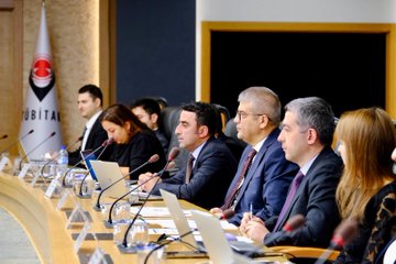 First image shows a long conference table in a modern room with wooden panels and flags including Turkish and Kosovo ones, where numerous professionals in suits sit on both sides engaged in a meeting, with laptops, documents, and a projector visible. Second image depicts a closer view of the meeting table with delegates including men and women in formal attire, microphones, water glasses, notebooks, and a Turkish flag in the background. Third image captures another segment of the table discussion with participants leaning forward, holding microphones, sipping from tea glasses, surrounded by papers and laptops. Fourth image features a large group of about 20 professionals standing in front of the TÜBİTAK building entrance with its logo and Turkish flag, dressed in business suits and dresses on steps with a blue carpet.