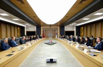 First image shows a long conference table in a modern room with wooden panels and flags including Turkish and Kosovo ones, where numerous professionals in suits sit on both sides engaged in a meeting, with laptops, documents, and a projector visible. Second image depicts a closer view of the meeting table with delegates including men and women in formal attire, microphones, water glasses, notebooks, and a Turkish flag in the background. Third image captures another segment of the table discussion with participants leaning forward, holding microphones, sipping from tea glasses, surrounded by papers and laptops. Fourth image features a large group of about 20 professionals standing in front of the TÜBİTAK building entrance with its logo and Turkish flag, dressed in business suits and dresses on steps with a blue carpet.