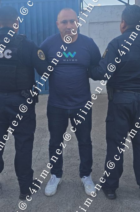 Three men stand in an outdoor area near a blue metal door and chain-link fence under an overcast sky. The central man wears a dark blue t-shirt with a white W logo, jeans, and white sneakers, facing forward with hands at sides. Two men in black tactical vests and pants, one with a badge on the arm, stand on either side, one holding the central mans arm. Logos including C4 and W appear on clothing.