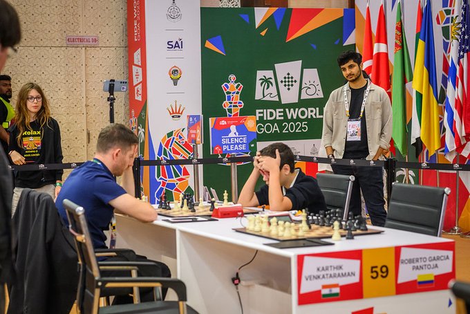 Chessboard setup with black and white pieces mid-game on a table between two players one in blue shirt focused on board and opponent in black shirt with hand on forehead tournament banners in background including FIDE World Cup 2025 logos flags from various countries SAI emblem and names Karthik Venkataraman and Robert Garca on score sheets audience members watching including man in light jacket and woman in yellow top silence sign visible.