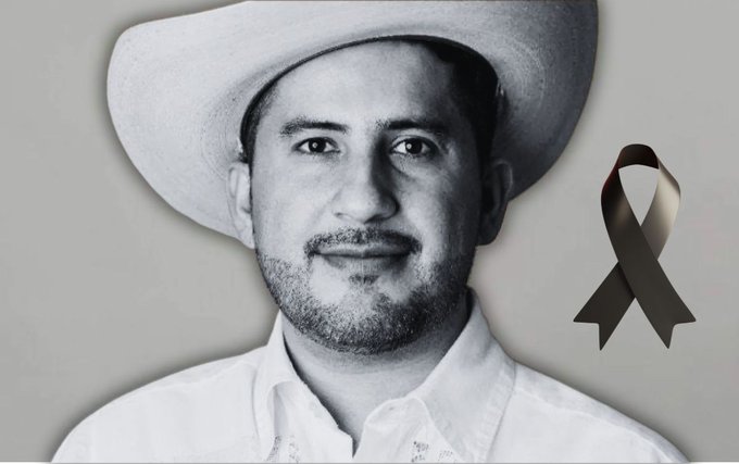 Black and white portrait of a man with short hair beard and mustache wearing a wide-brimmed cowboy hat and white collared shirt against a gray background with a black ribbon in the lower right corner symbolizing mourning.