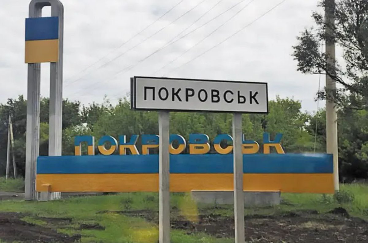 路标以黑色背景显示 ПОКРОВСК，安装在白色柱子上，旁边是蓝色和黄色标志，黄色背景蓝色字母 ПОКРОВСК，由金属杆支撑，背景是绿色树木、灰色天空和电线杆