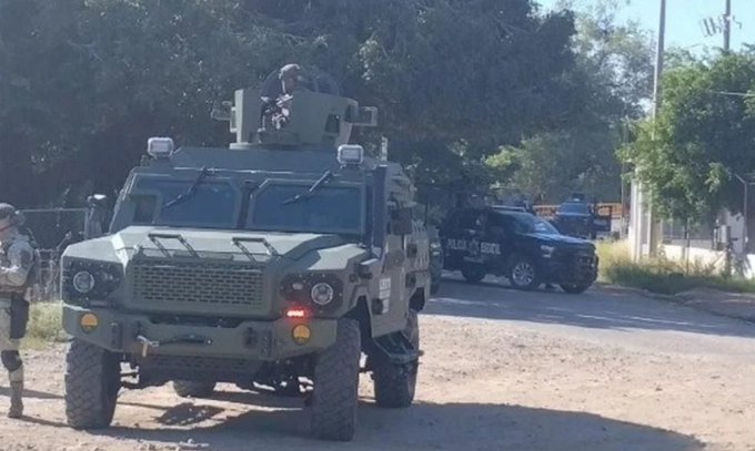 Green armored military vehicle with mounted turret and personnel in tactical gear positioned on a dirt road beside trees and a metal fence another black SUV in background under clear sky with utility poles visible