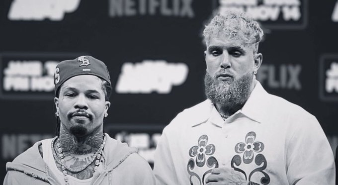 Black and white photo of two men standing side by side against a backdrop with Netflix and Most Valuable Promotions logos. Left man wears a red San Francisco Giants cap, gold chain necklace, and casual shirt. Right man has curly blond hair, beard, and wears a white shirt with floral designs.