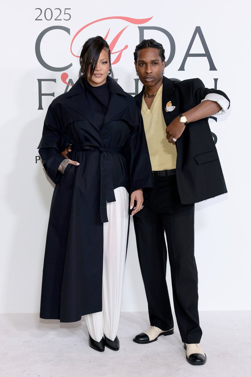 Rihanna stands wearing a long black coat with belt over white pants and black heels, her hair styled in a ponytail, posing with hand on hip. A$AP Rocky beside her wears a yellow shirt under beige jacket, gold chain, watch, black pants, and white shoes with black accents. They stand together smiling at the 2025 CFDA Fashion Awards backdrop featuring the CFDA logo.