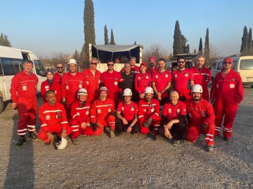 First image shows six men in red firefighting suits and white helmets standing on a truck bed lifting a yellow metal ladder near a gray building with glass windows and a metal scaffold under a clear blue sky with pine trees. Second image depicts a large group of about 20 people in red suits some wearing helmets posing together on gravel ground near white vans and tents with pine trees and a clear sky in the background. Third image features four men in red suits and helmets standing near concrete blocks holding a red fire extinguisher with construction materials and trees visible. Fourth image displays several white and blue tents set up on gravel with a few men in red suits walking around under a sunny sky with setting sun rays and pine trees.