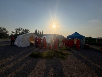 First image shows six men in red firefighting suits and white helmets standing on a truck bed lifting a yellow metal ladder near a gray building with glass windows and a metal scaffold under a clear blue sky with pine trees. Second image depicts a large group of about 20 people in red suits some wearing helmets posing together on gravel ground near white vans and tents with pine trees and a clear sky in the background. Third image features four men in red suits and helmets standing near concrete blocks holding a red fire extinguisher with construction materials and trees visible. Fourth image displays several white and blue tents set up on gravel with a few men in red suits walking around under a sunny sky with setting sun rays and pine trees.