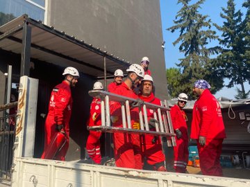 First image shows six men in red firefighting suits and white helmets standing on a truck bed lifting a yellow metal ladder near a gray building with glass windows and a metal scaffold under a clear blue sky with pine trees. Second image depicts a large group of about 20 people in red suits some wearing helmets posing together on gravel ground near white vans and tents with pine trees and a clear sky in the background. Third image features four men in red suits and helmets standing near concrete blocks holding a red fire extinguisher with construction materials and trees visible. Fourth image displays several white and blue tents set up on gravel with a few men in red suits walking around under a sunny sky with setting sun rays and pine trees.