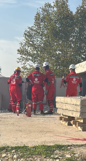 First image shows six men in red firefighting suits and white helmets standing on a truck bed lifting a yellow metal ladder near a gray building with glass windows and a metal scaffold under a clear blue sky with pine trees. Second image depicts a large group of about 20 people in red suits some wearing helmets posing together on gravel ground near white vans and tents with pine trees and a clear sky in the background. Third image features four men in red suits and helmets standing near concrete blocks holding a red fire extinguisher with construction materials and trees visible. Fourth image displays several white and blue tents set up on gravel with a few men in red suits walking around under a sunny sky with setting sun rays and pine trees.