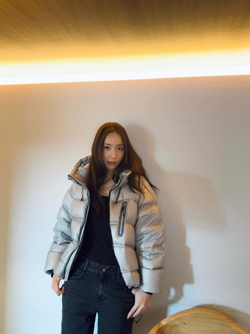 First image shows a young Asian woman with shoulder-length brown hair wearing a light gray hooded puffer jacket over a black top and dark pants sitting on a wooden chair at a wooden table holding a small object in her hands with a neutral expression in a plain room with beige walls. Second image depicts the same woman standing against a beige wall with her arms crossed wearing the same light gray puffer jacket black top and dark pants with long brown hair loose and a slight smile in an indoor setting with warm yellow lighting and wooden furniture nearby.