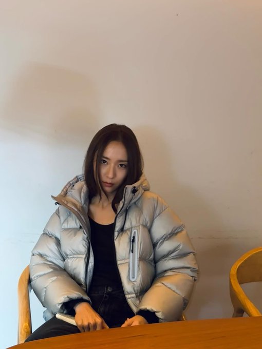 First image shows a young Asian woman with shoulder-length brown hair wearing a light gray hooded puffer jacket over a black top and dark pants sitting on a wooden chair at a wooden table holding a small object in her hands with a neutral expression in a plain room with beige walls. Second image depicts the same woman standing against a beige wall with her arms crossed wearing the same light gray puffer jacket black top and dark pants with long brown hair loose and a slight smile in an indoor setting with warm yellow lighting and wooden furniture nearby.
