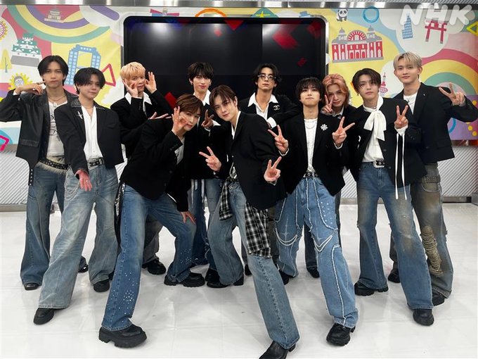 Eleven young men identified as INI group members stand and pose dynamically in a studio setting. They wear coordinated black and white outfits including jackets shirts and jeans with some plaid elements. Several make peace sign gestures while others smile or look at camera. Background features vibrant colorful abstract wall art and a large black screen displaying red and white Japanese text あたらしい above multicolored graphics.