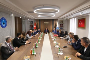 First image shows a large group of about 20 professionals in business suits standing in a circle in a modern conference room with curved wooden walls, ceiling lights, and a red accent wall. Second image depicts a long conference table with men and women in formal attire seated around it, Turkish and union flags on walls, bottled drinks and notepads on the table, under circular ceiling lights. Third image features four men in suits at a table with Turkish flags, plates of nuts, and beverages, in a room with wooden shelves and Kamu sign. Fourth image shows a similar meeting setup with a group around a long table, Turkish flags, union emblems, drinks, and snacks, in a formal office space.