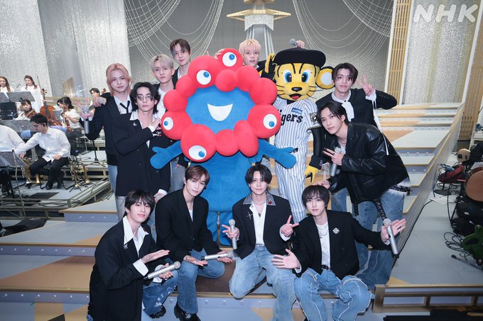 Group of young male performers in black suits and casual outfits posing on stage with blue smiling mascot character having red tentacles and yellow tiger mascot wearing blue jersey next to them surrounded by musical instruments orchestra background and decorative elements like stars and curtains.