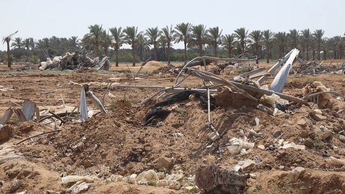 Aerial view of vast barren sandy field with scattered twisted metal debris resembling destroyed aircraft wreckage and building remnants amid dirt mounds and scattered rocks under clear sky with distant line of green palm trees on horizon.