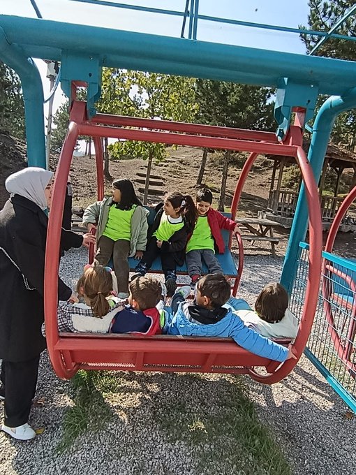 Group of young children in colorful vests and casual clothes stand smiling on wooden steps under a covered pavilion with trees and blue sky in background several adults in headscarves and casual attire pose with them. Children in vests sit at wooden tables drawing with crayons and colored pencils on paper surrounded by art supplies and books one table has a blackboard easel labeled Orman Okulu 2025. Children in vests draw at another wooden table with crayons pencils and books blackboard shows Orman Okulu 2025. Group of young children in vests sit in red seats of a blue cable car playground structure with trees and benches in background two adults in headscarves stand nearby.