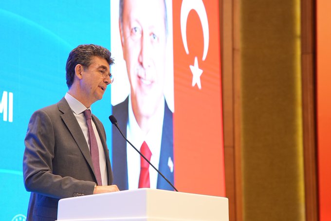 A man in a suit and tie stands at a podium speaking into a microphone, with a large screen behind him displaying a portrait of Recep Tayyip Erdogan and Turkish flags. The setting is a formal conference room with blue and orange lighting. Another image shows a wide view of the event stage with banners reading 2024 YLSY Bursiyerleri Vizyon ve Farkındalık Eğitimi, flanked by Turkish flags, speakers on stage, and an audience seated in chairs below.