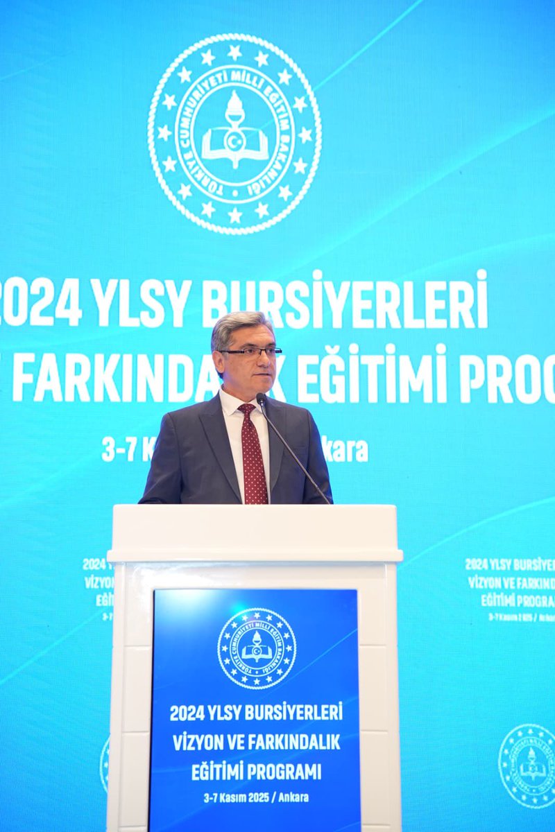 A man in a suit and tie stands at a podium speaking into a microphone during an event. Behind him is a large screen displaying the text 2024 YLSY Bursiyerleri Farkındalık ve Vizyon Eğitimi Programı 3-7 Kasım Ankara in Turkish along with the emblem of the Ministry of National Education. The backdrop features the Turkish flag and the ministry logo on a blue background. The setting appears to be a formal conference room.