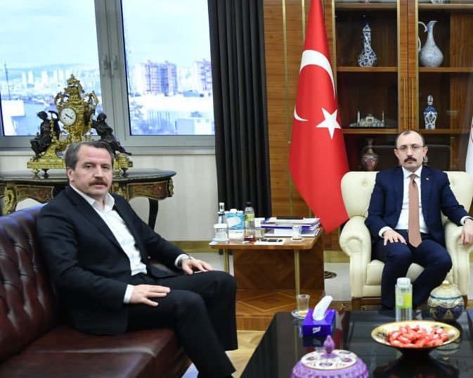 First image shows three men in dark suits and ties standing formally in an office with wooden floors and paneling. Behind them are framed portraits of Mustafa Kemal Atatürk on the walls, Turkish flags on poles, a yellow-starred banner, red curtains, a green plant, and a desk with stacked white binders and a vase. The men appear to be middle-aged with short hair, one with a mustache, positioned side by side. Second image depicts two men in suits seated on a brown leather couch in a luxurious room with large windows overlooking a cityscape of buildings. A Turkish flag stands nearby, ornate wooden furniture including a clock and vases with decor, a low table with water bottles, stacked papers, white tissues, and a plate of strawberries.
