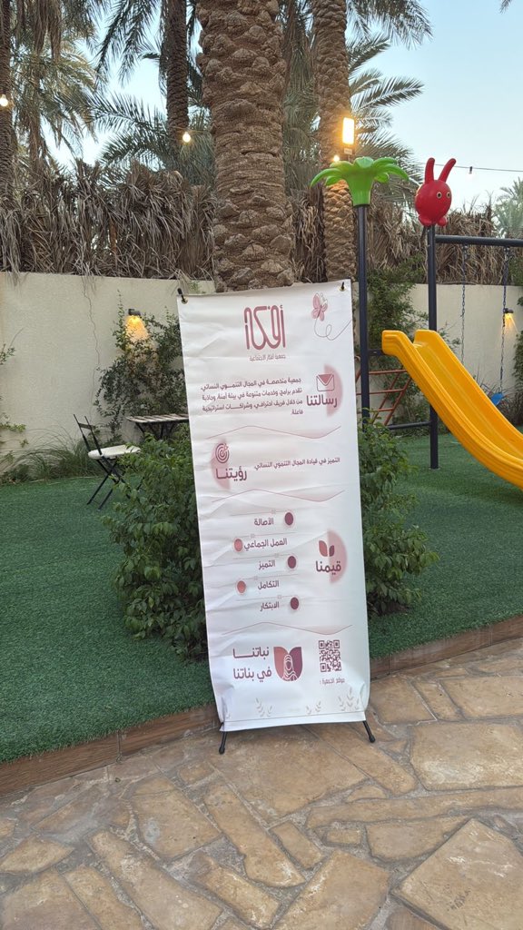 First image displays a vase of white flowers with green stems on a wooden table next to a pink card from the association with contact details and Arabic text, and a white framed plaque with Arabic inscription. Second image shows an outdoor courtyard with palm trees, string lights, a red balloon palm tree decoration, a yellow slide on green grass, chairs, and a tall white banner with red Arabic text about the event including a QR code.
